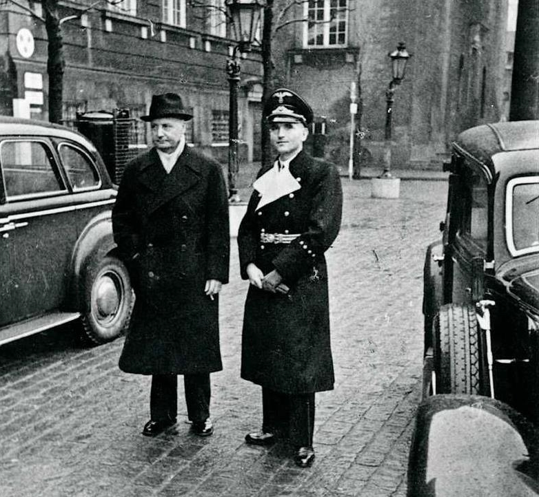 Fotografiet viser Erik Scavenius, Danmarks statsminister sammen med Dr. Werner Best, repræsentant for den tyske besættelsesmagt. Billedet er taget den 6. februar 1943 da Werner Best blev præsenteret for den danske kronprins efter at han i sine første måne